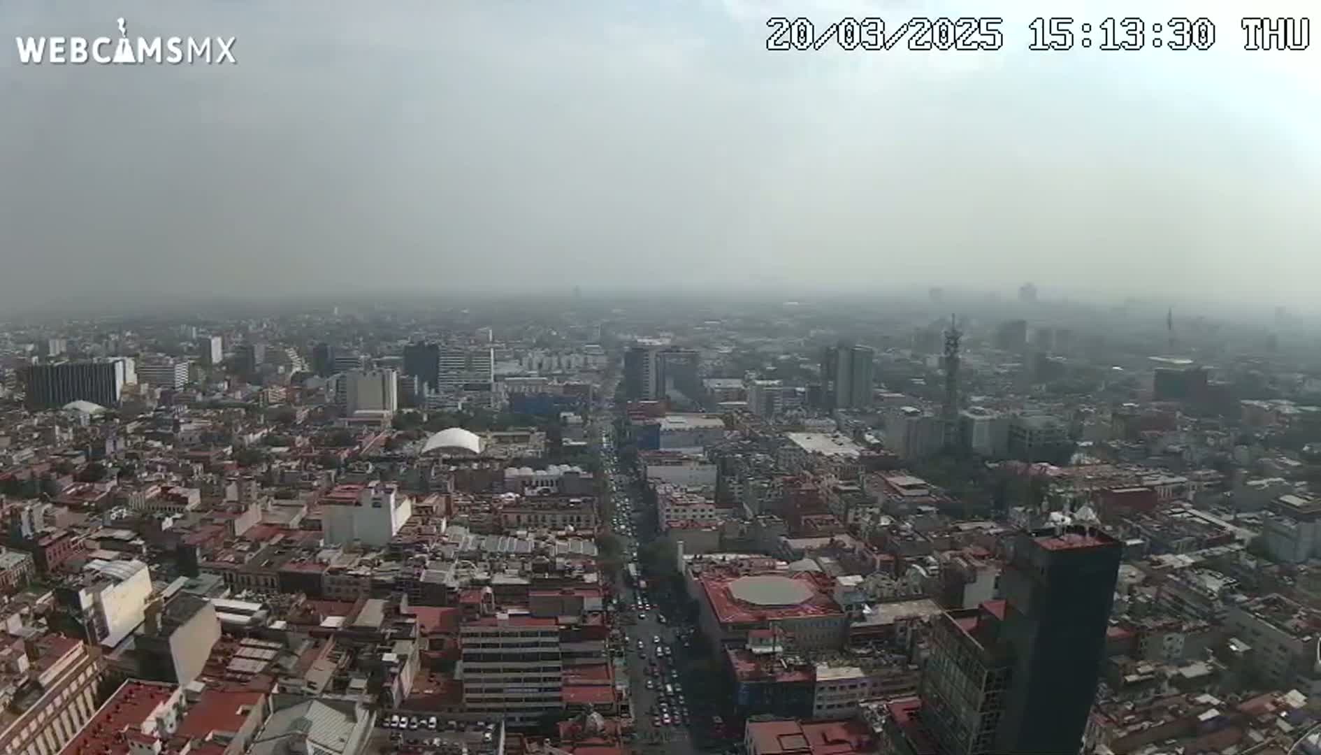 A hazy, high-angle view shows a dense cityscape extending to the horizon under a mostly cloudy sky.