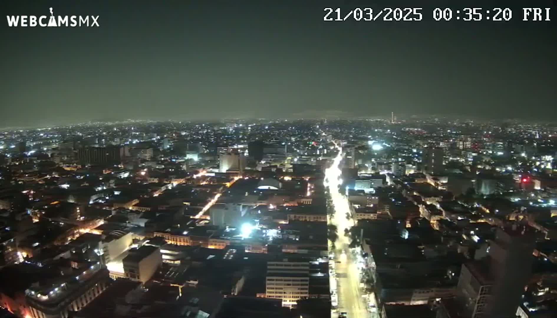A nighttime aerial view of a city shows a brightly lit main road cutting through a sprawling network of darker streets and buildings under a hazy sky.