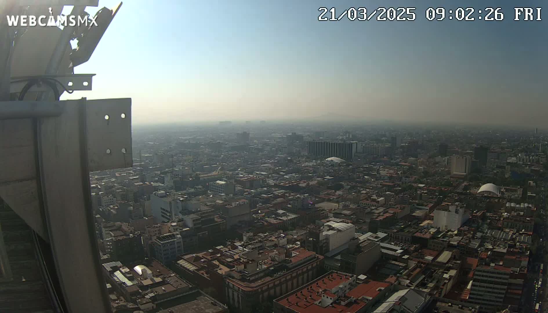A hazy, sunny view from a tall structure shows a vast expanse of densely packed city buildings stretching to the horizon.