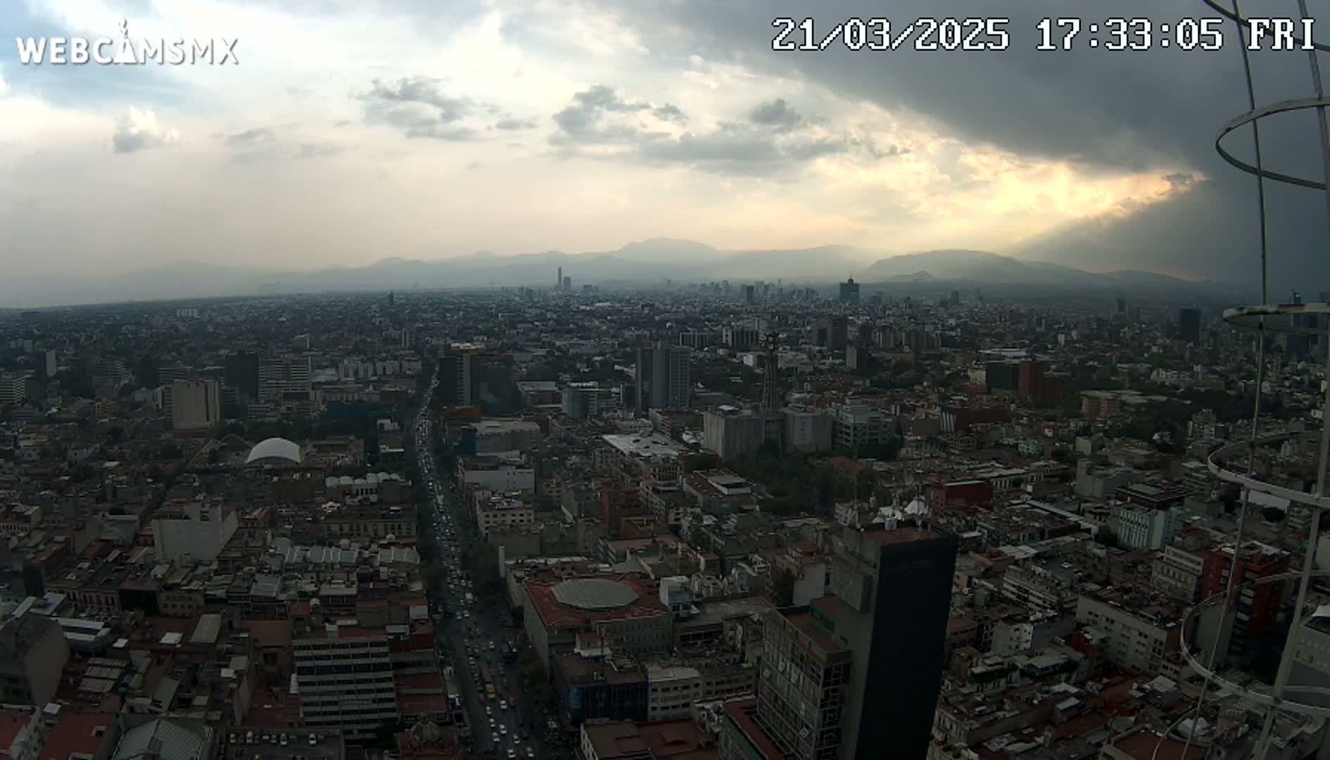 An aerial view of a large city under a partly cloudy sky shows distant mountains in the background.