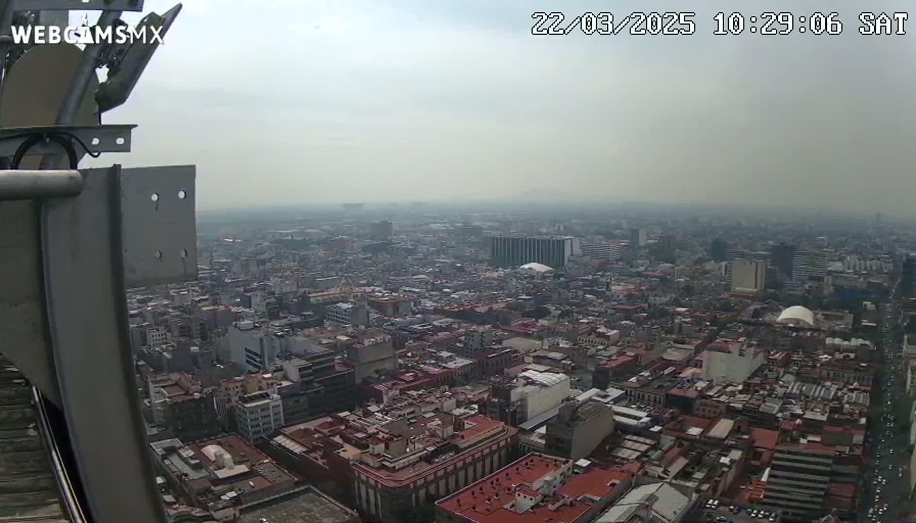 A hazy, high-angle view shows a sprawling city with many closely packed buildings and red-tiled roofs, seen from a tall structure.