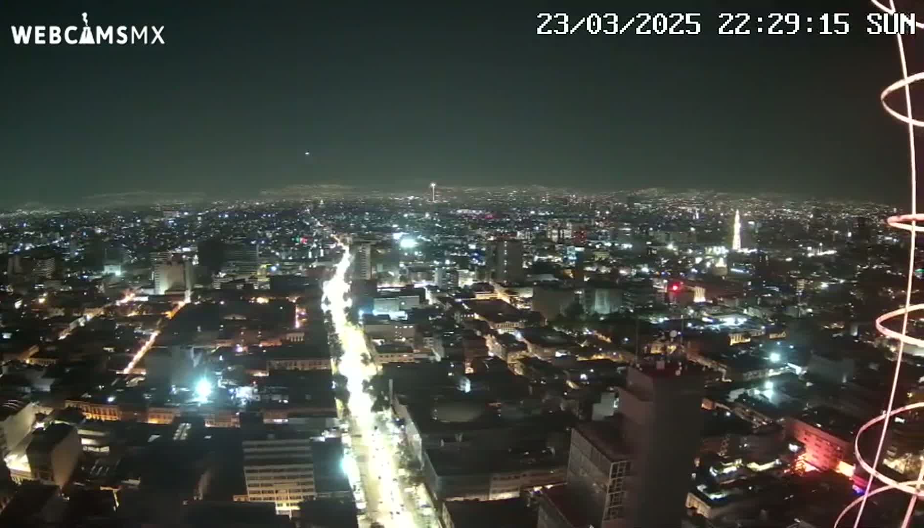 A nighttime, aerial view shows a sprawling city illuminated by countless lights, with a long, brightly lit street cutting through the urban landscape.
