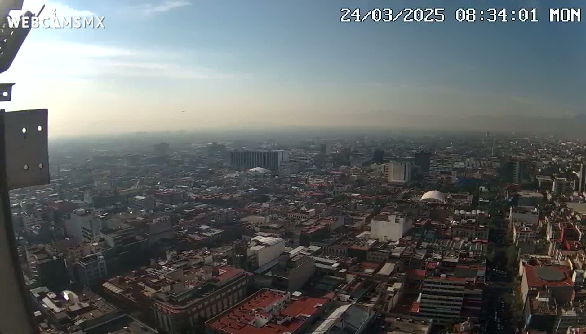A hazy, high-angle view shows a sprawling city with numerous buildings under a mostly clear sky.