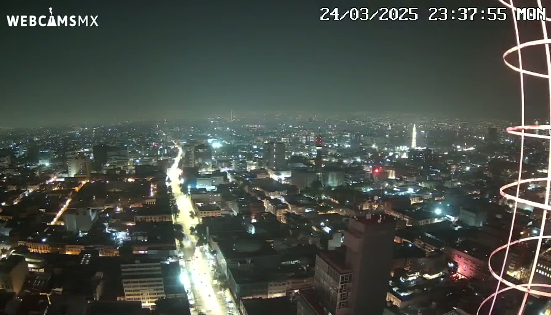 A nighttime aerial view of a sprawling city with brightly lit streets and buildings under a hazy sky.