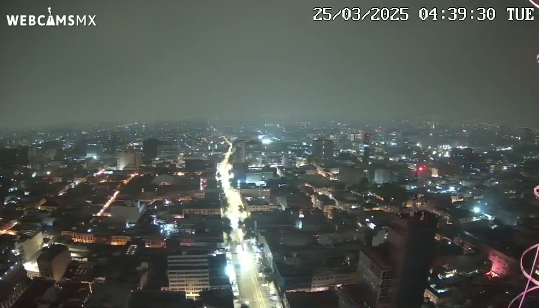 A nighttime aerial view of a city with hazy or foggy conditions shows a long, brightly lit street cutting through a densely packed urban area.