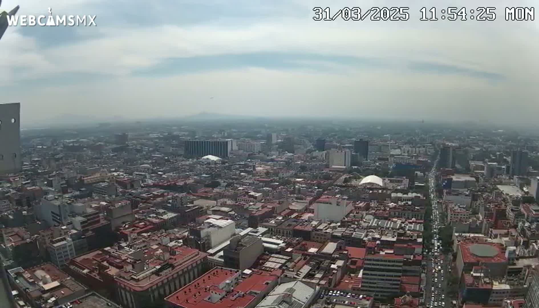A hazy, high-angle view shows a sprawling city with numerous buildings and streets, under a mostly cloudy sky.