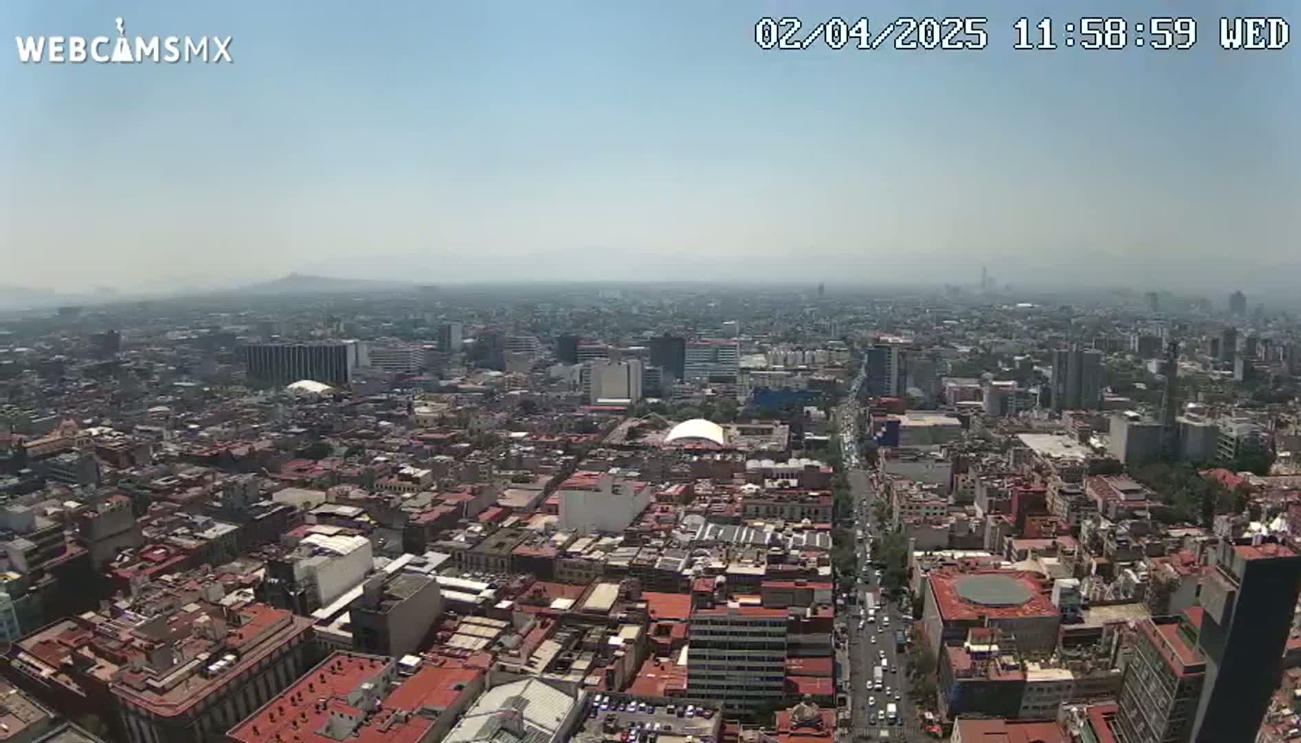 A hazy, sunny day overlooks a sprawling cityscape of densely packed buildings, with a long road visible cutting through the urban expanse.