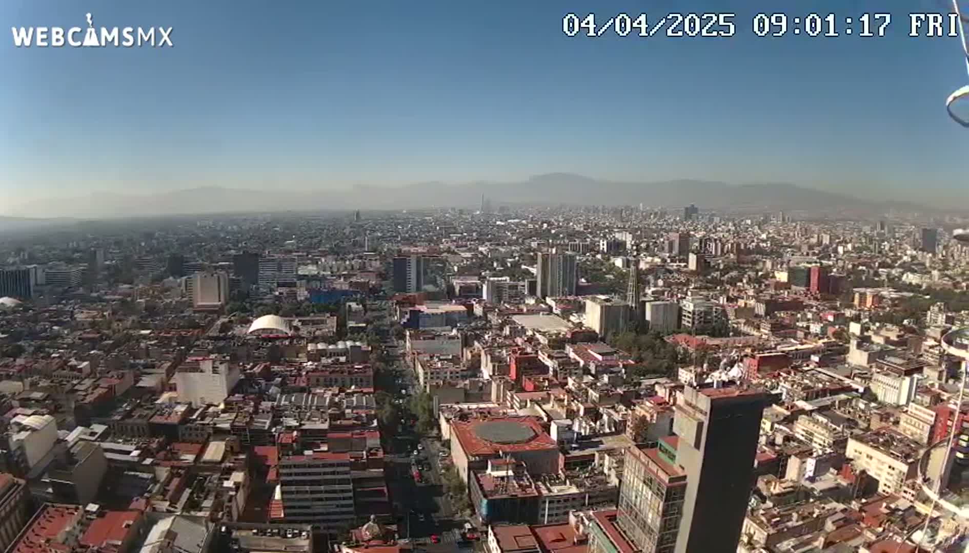 A high-angle, long shot of a sprawling city on a clear, sunny day with distant mountains visible on the horizon.