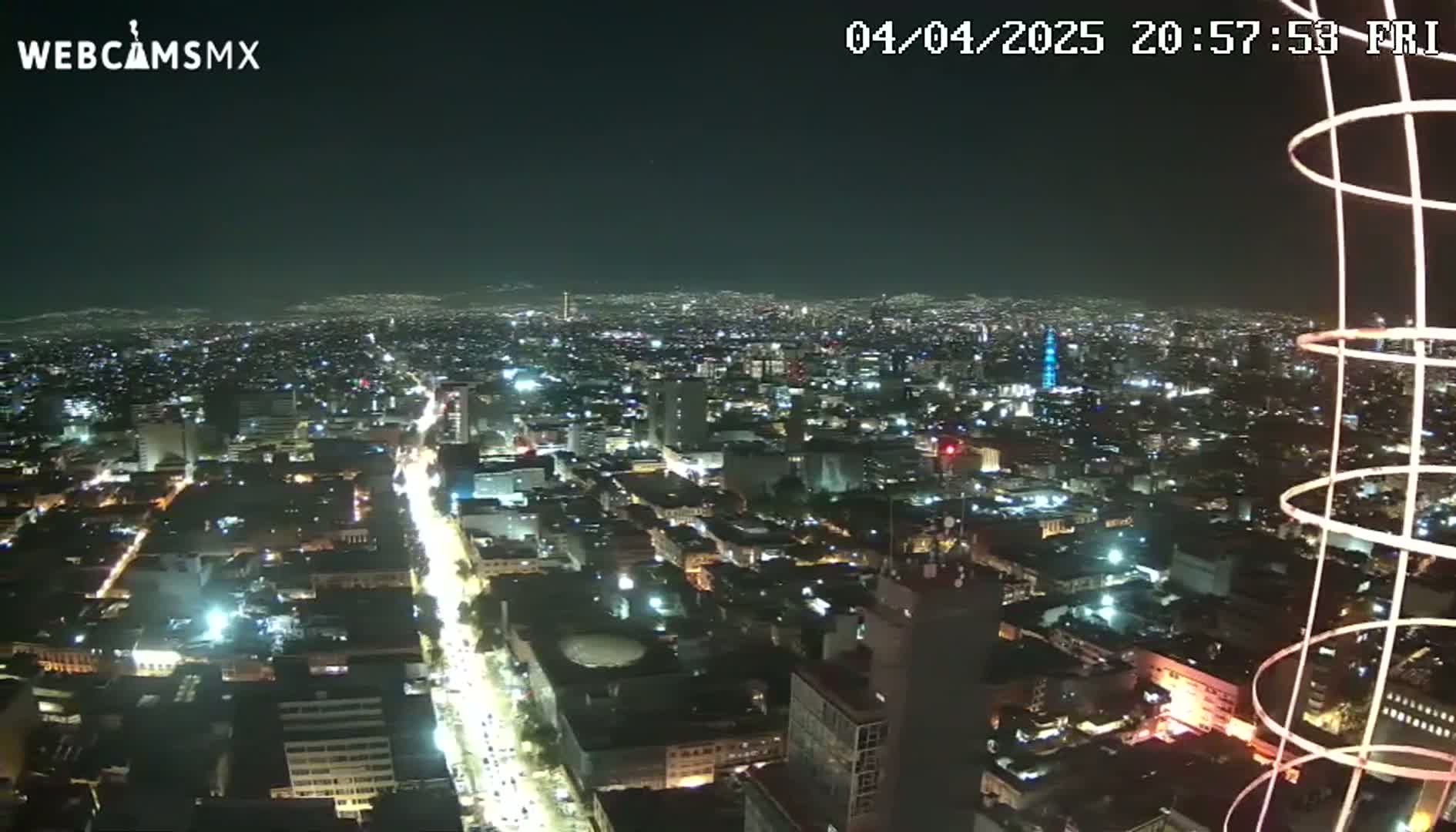 A nighttime, high-angle view of a sprawling city illuminated by numerous lights, with a portion of a tall structure visible in the lower right corner.