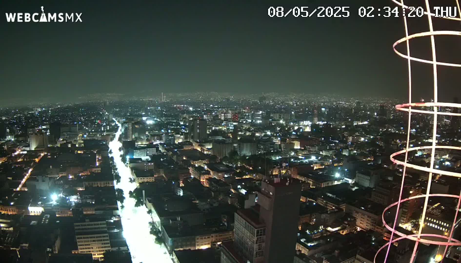 A nighttime aerial view of a brightly lit city, with a long, illuminated street visible, and a portion of a tall, circular structure in the foreground.