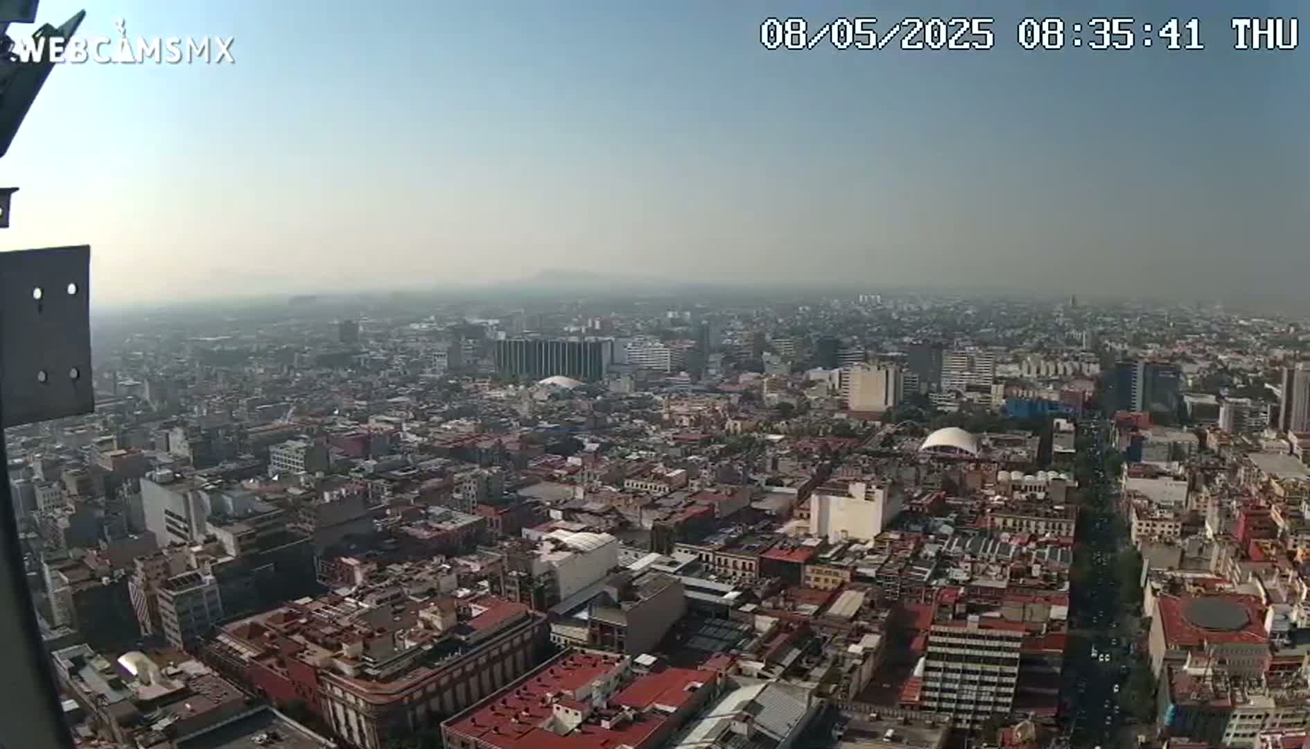 A hazy, high-angle view of a sprawling city on a clear day shows a dense collection of buildings extending to the horizon, with a mountain visible in the far distance.