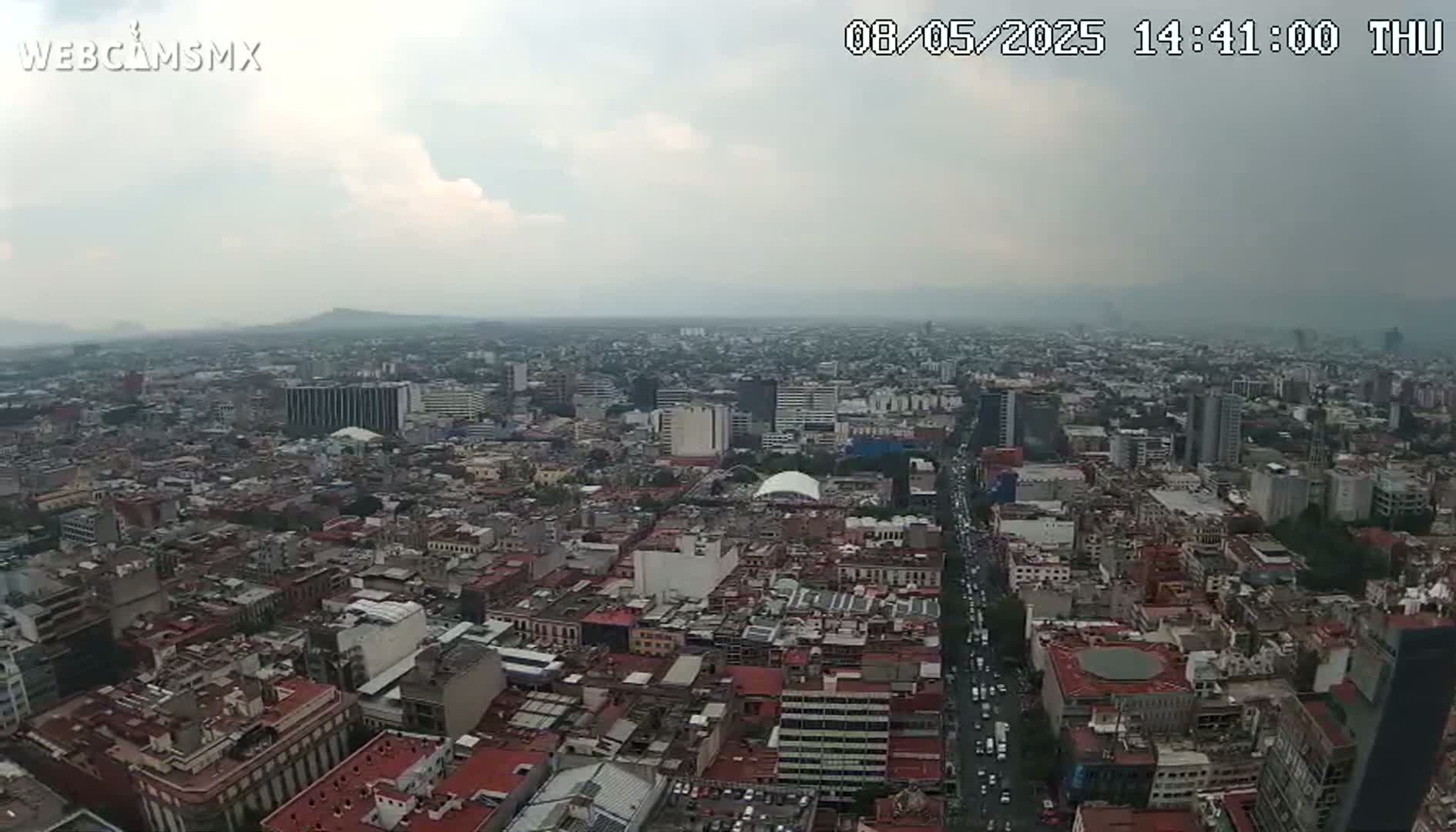 A sprawling cityscape under an overcast sky is viewed from a high vantage point, showing numerous buildings and streets stretching to distant mountains.