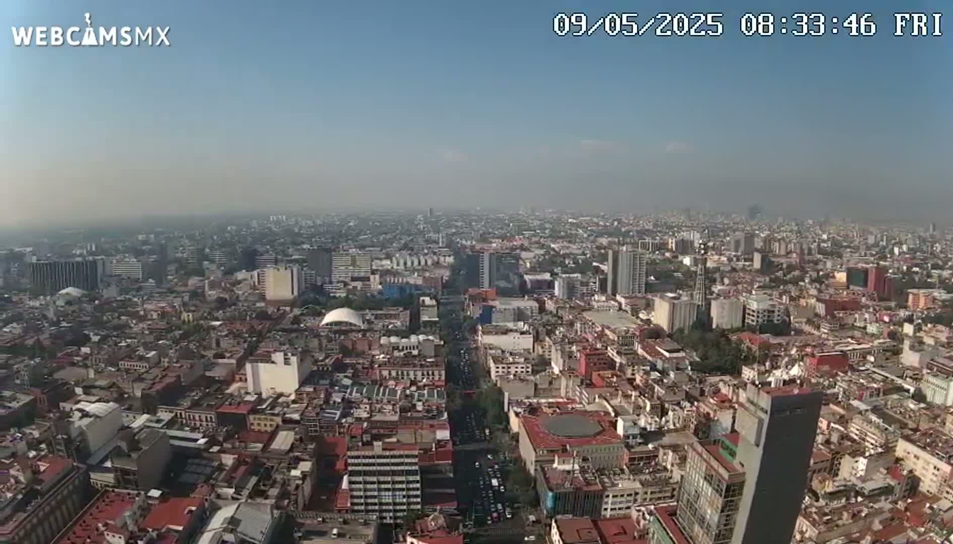 A hazy, sunny day reveals a sprawling cityscape, viewed from a high vantage point, showing densely packed buildings and a wide avenue with traffic.