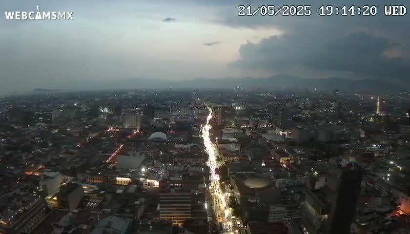 A nighttime, high-angle view of a sprawling city under a cloudy sky shows a long, illuminated road cutting through the urban landscape.