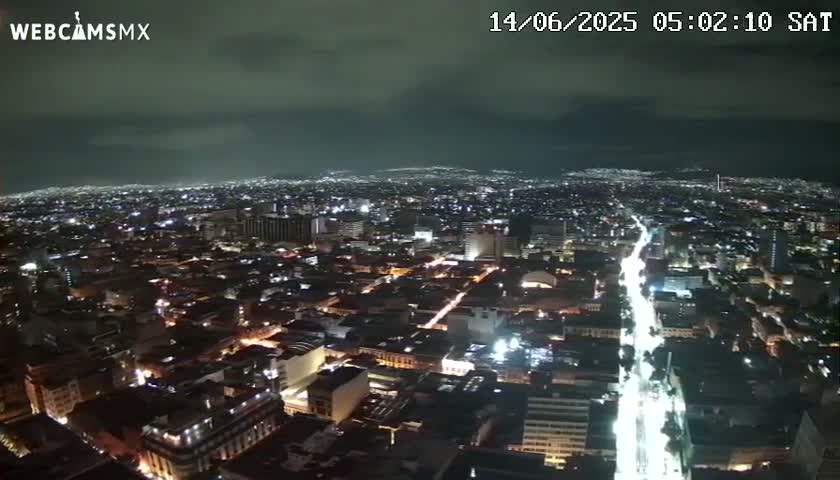 A nighttime, high-angle view of a sprawling city illuminated by streetlights and building lights under a mostly cloudy sky.