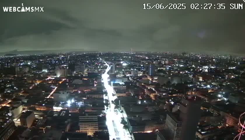 A nighttime aerial view of a sprawling city under a cloudy sky shows a long, brightly lit street cutting through the urban landscape.