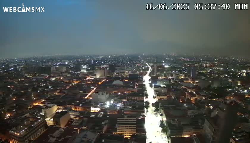 A nighttime, high-angle view of a city with a brightly lit street extending from the foreground into the distance under a hazy sky.