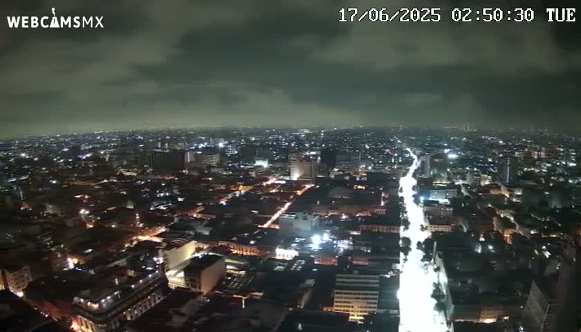 A nighttime aerial view of a sprawling city under a cloudy sky shows a brightly lit street cutting through the darker urban landscape.