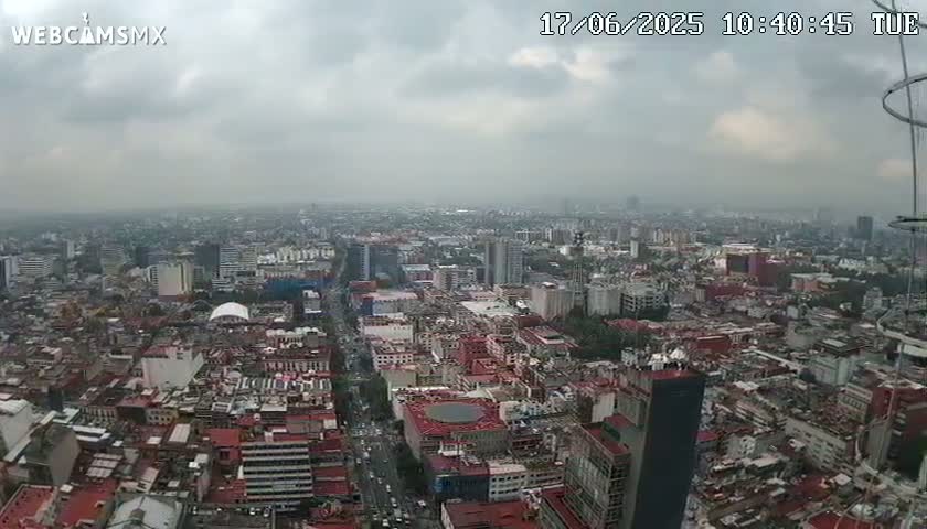 A high-angle, long shot of a sprawling city under an overcast sky shows mostly low-rise buildings with some high-rises scattered throughout.
