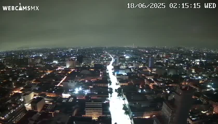 A nighttime aerial view shows a brightly lit city street cutting through a sprawling metropolis under a cloudy sky.
