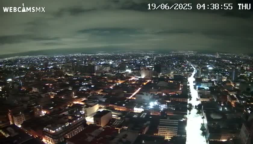 A nighttime aerial view of a sprawling city with overcast skies shows a brightly lit street cutting through the urban landscape.