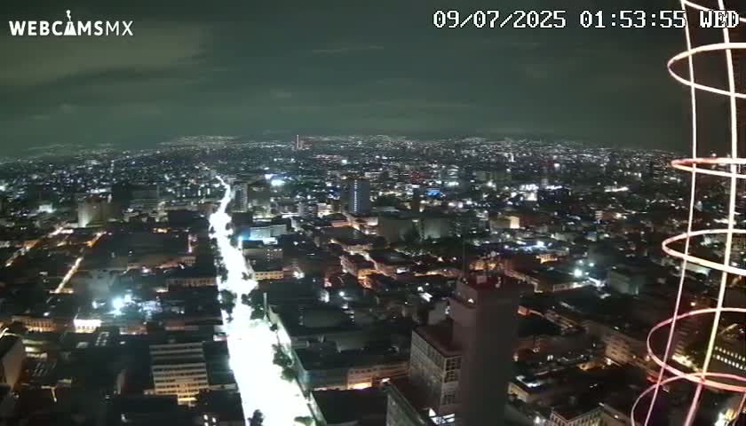 A nighttime, high-angle view of a sprawling city with brightly lit streets and buildings, partially obscured by a metallic structure on the right.
