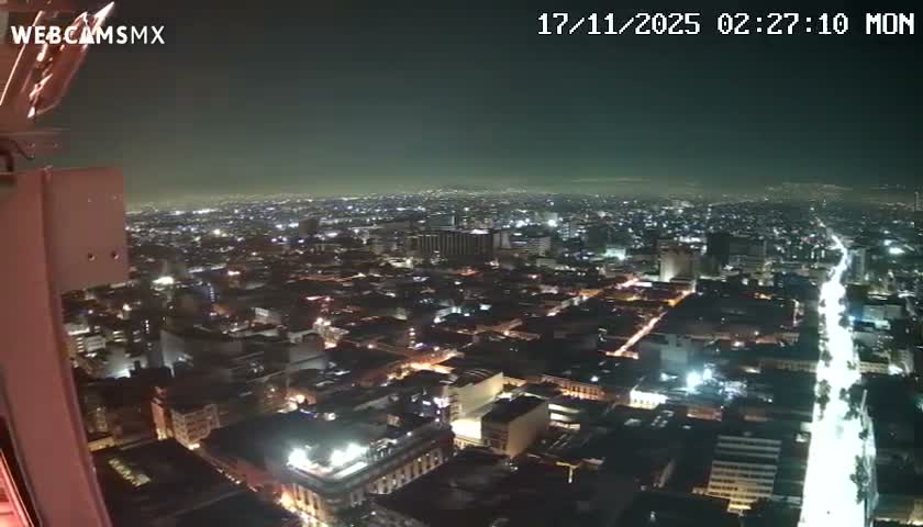 An expansive aerial view reveals a city at night, showcasing a dense grid of illuminated buildings and a brightly lit main thoroughfare stretching into the distance under a clear, dark sky.