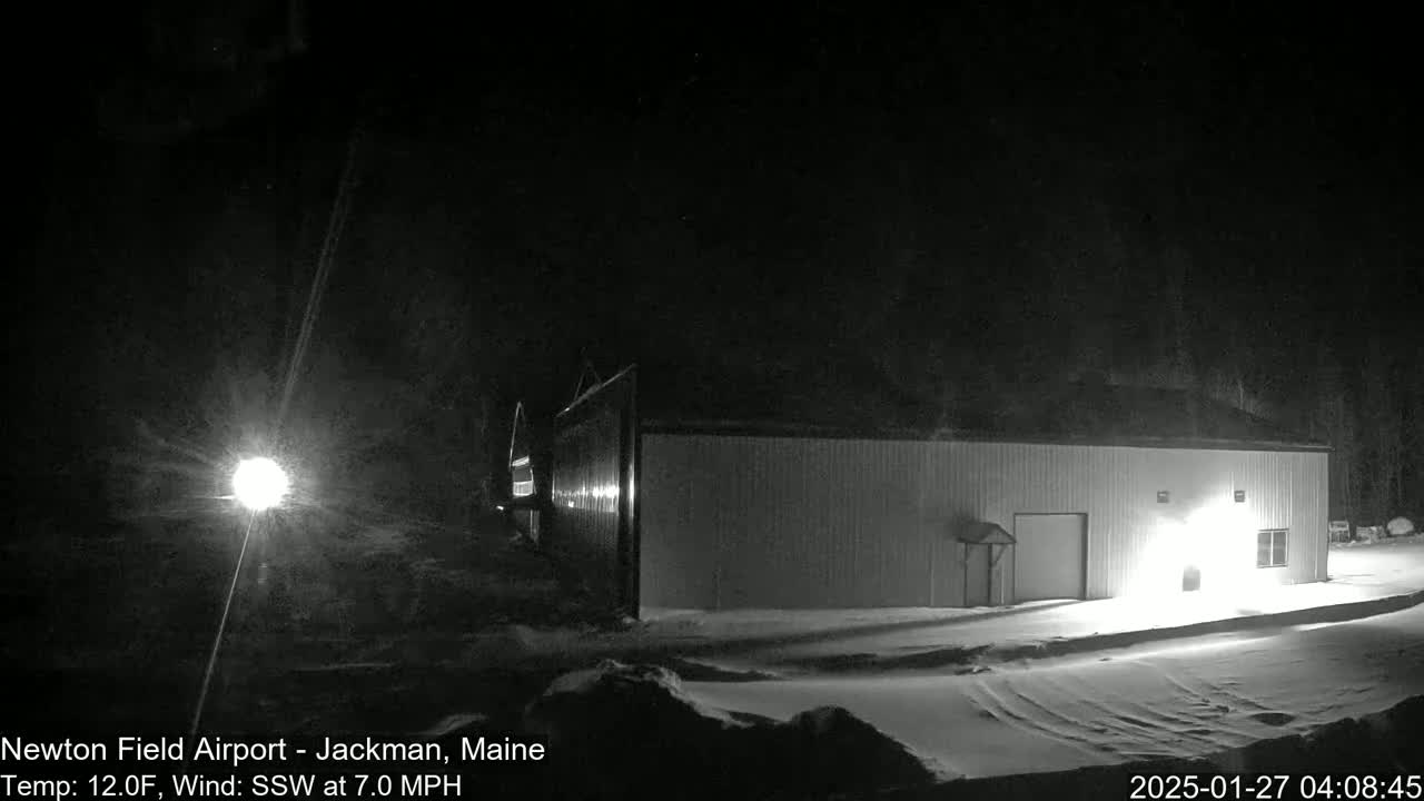 A long, light-colored building is visible under a dark, nighttime sky with snow on the ground, and a light source illuminates the scene.