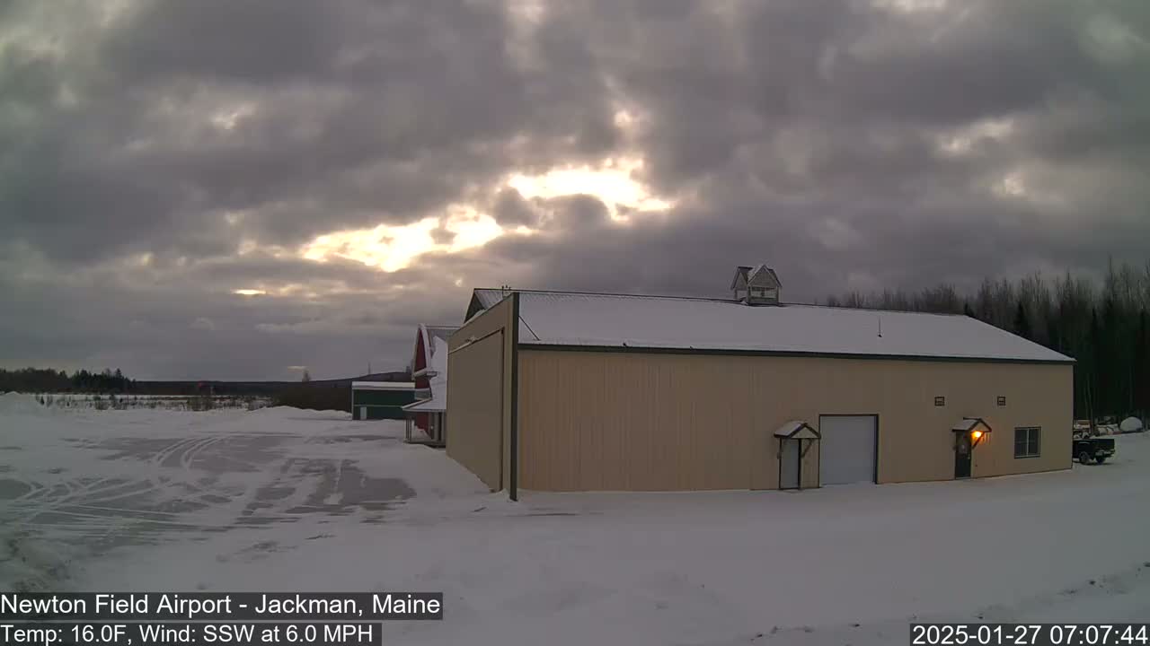 A tan hangar sits in a snow-covered field under a cloudy sky with some sunlight breaking through.