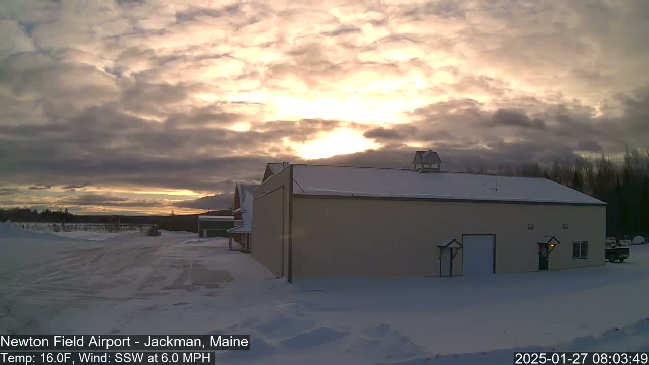 A large tan building with a snow-covered roof sits in a snowy landscape under a partly cloudy sunrise.