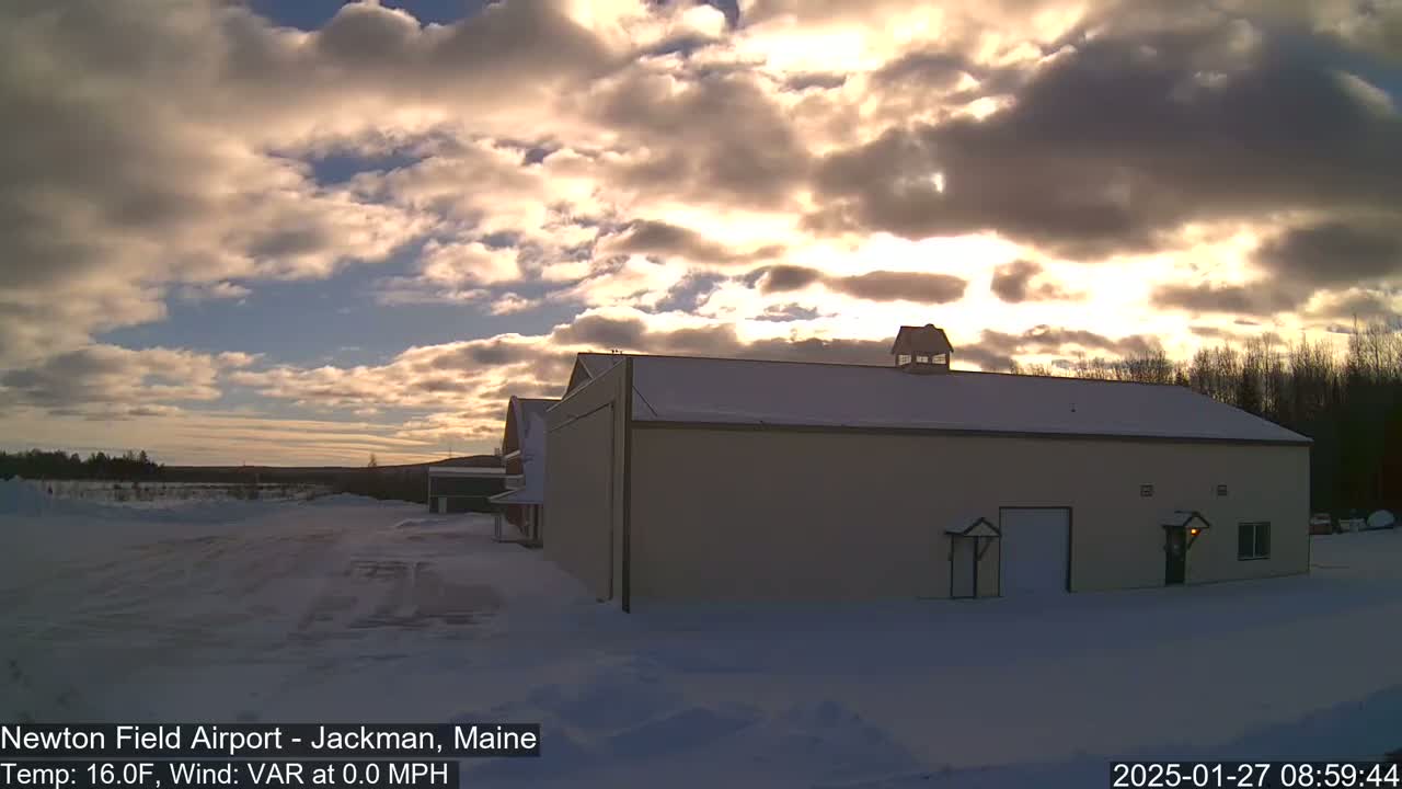 A tan building with a snow-covered roof stands in a snow-covered landscape under a partly cloudy sky at sunset.