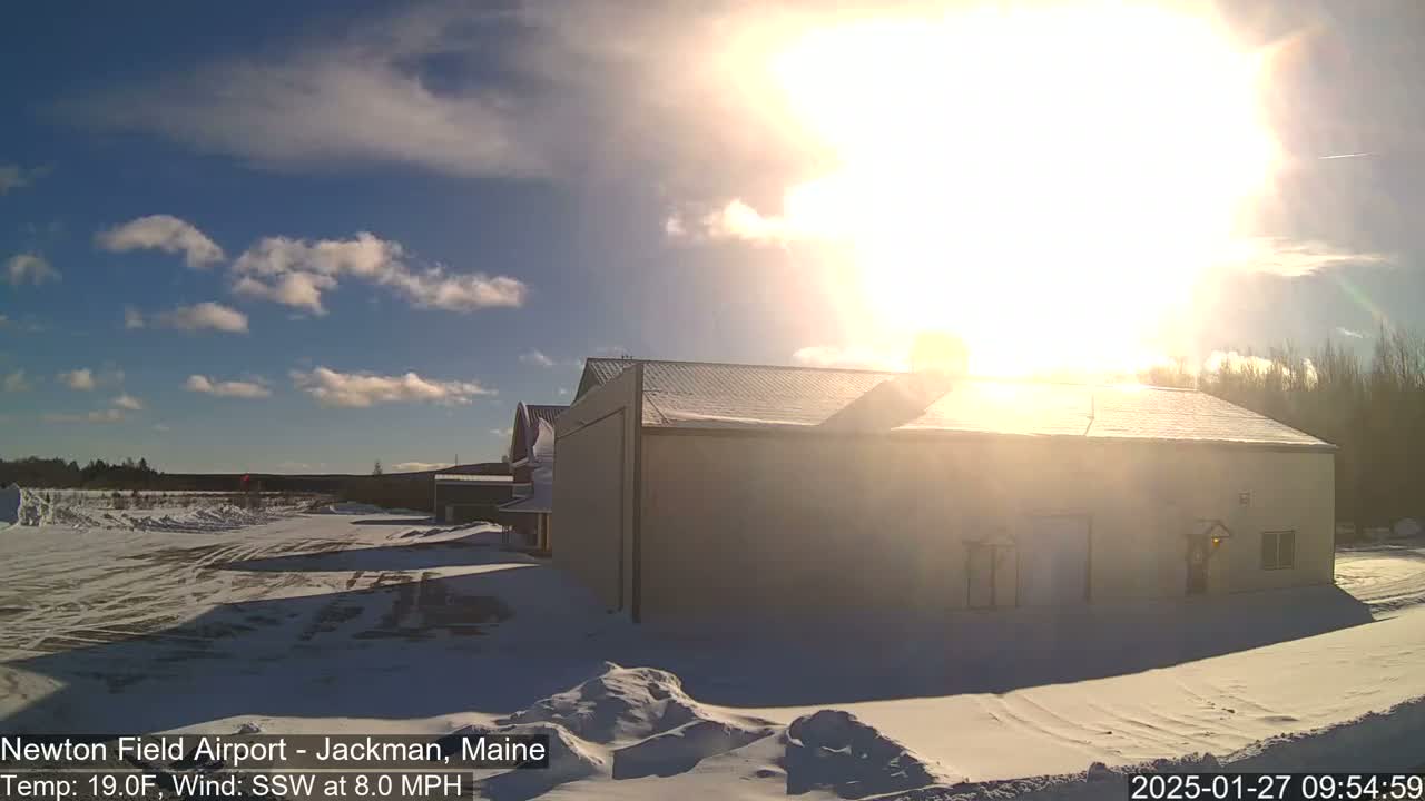 A snow-covered airfield with a large building in the foreground, bathed in bright sunlight under a partly cloudy sky.