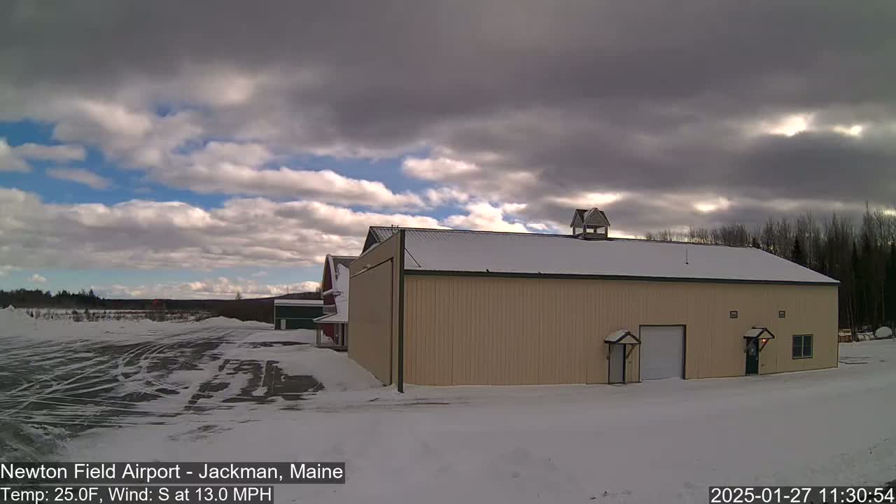 A tan hangar sits in a snow-covered airfield under a mostly cloudy sky with some blue showing through.
