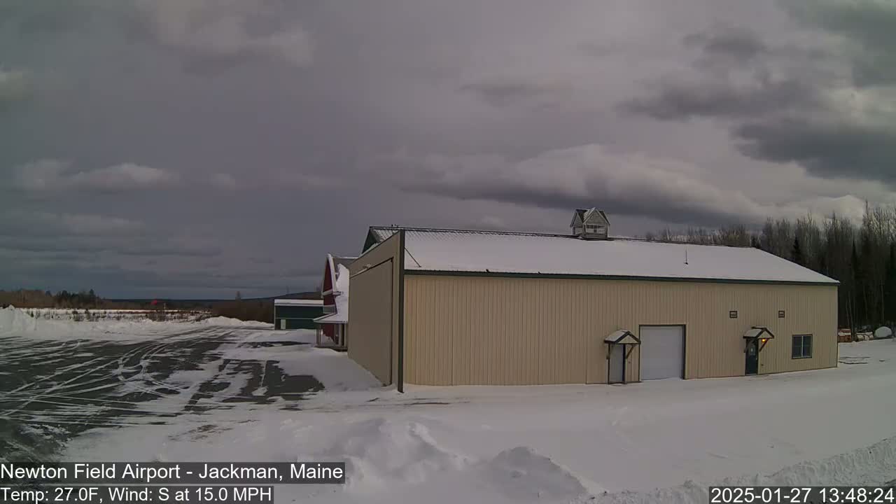 A tan hangar sits in a snow-covered airfield under a cloudy sky with a 27°F temperature and a 15 mph south wind.