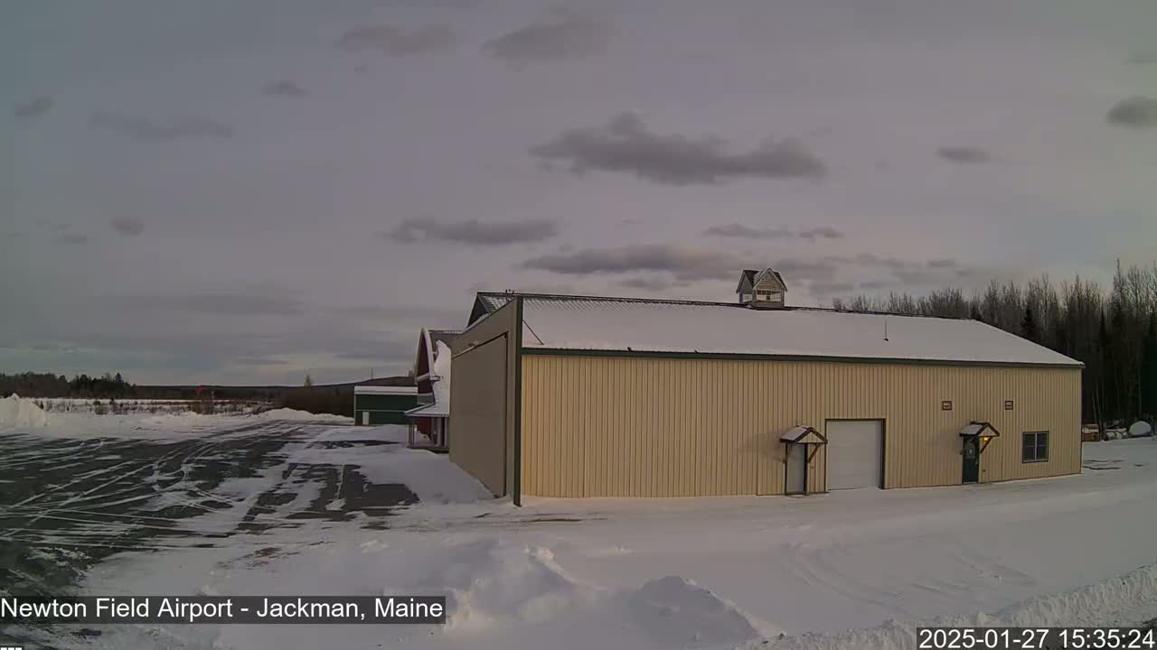 A large, light beige hangar-like building sits in a snow-covered airfield under a cloudy, overcast sky.