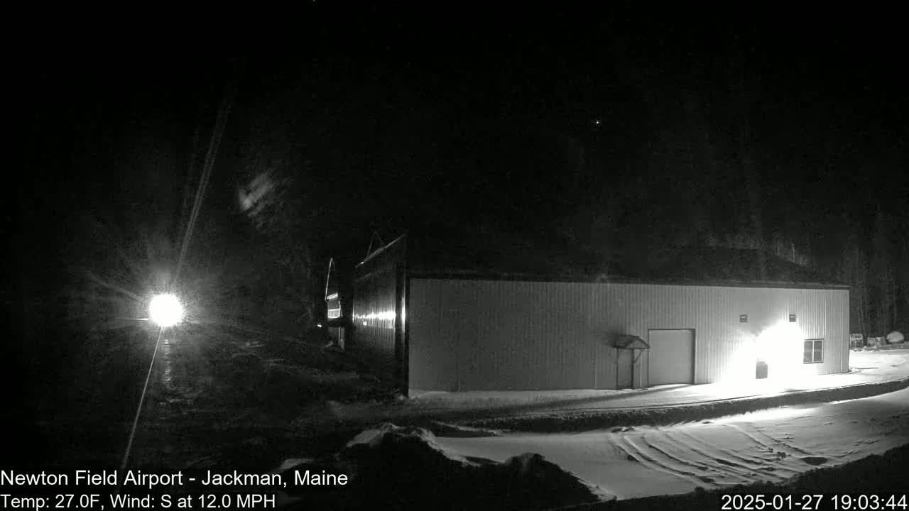 A long, light-colored building is illuminated at night in snowy conditions with a south wind of 12 mph.