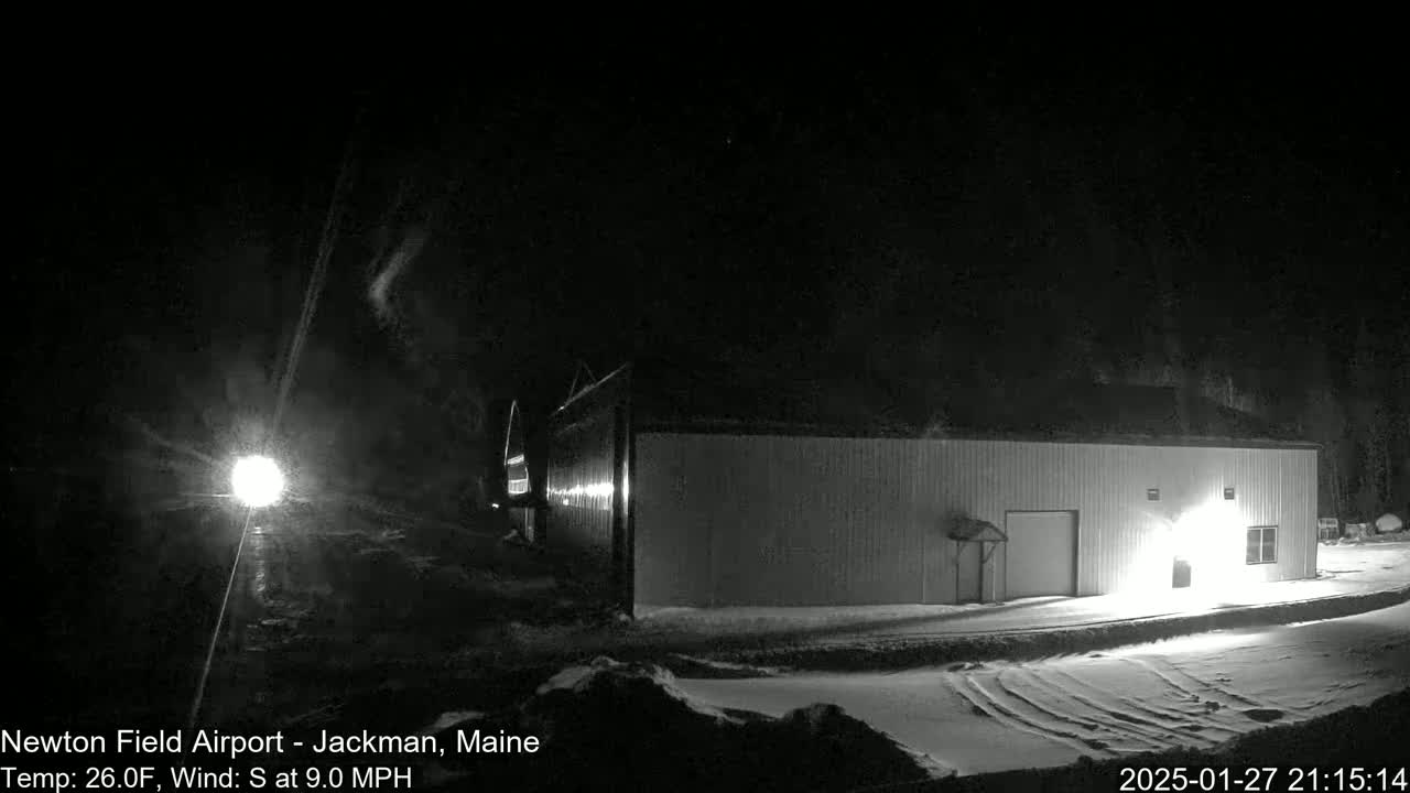 A long, light-grey building is illuminated at night in snowy conditions with a south wind at 9.0 mph and a temperature of 26.0°F.
