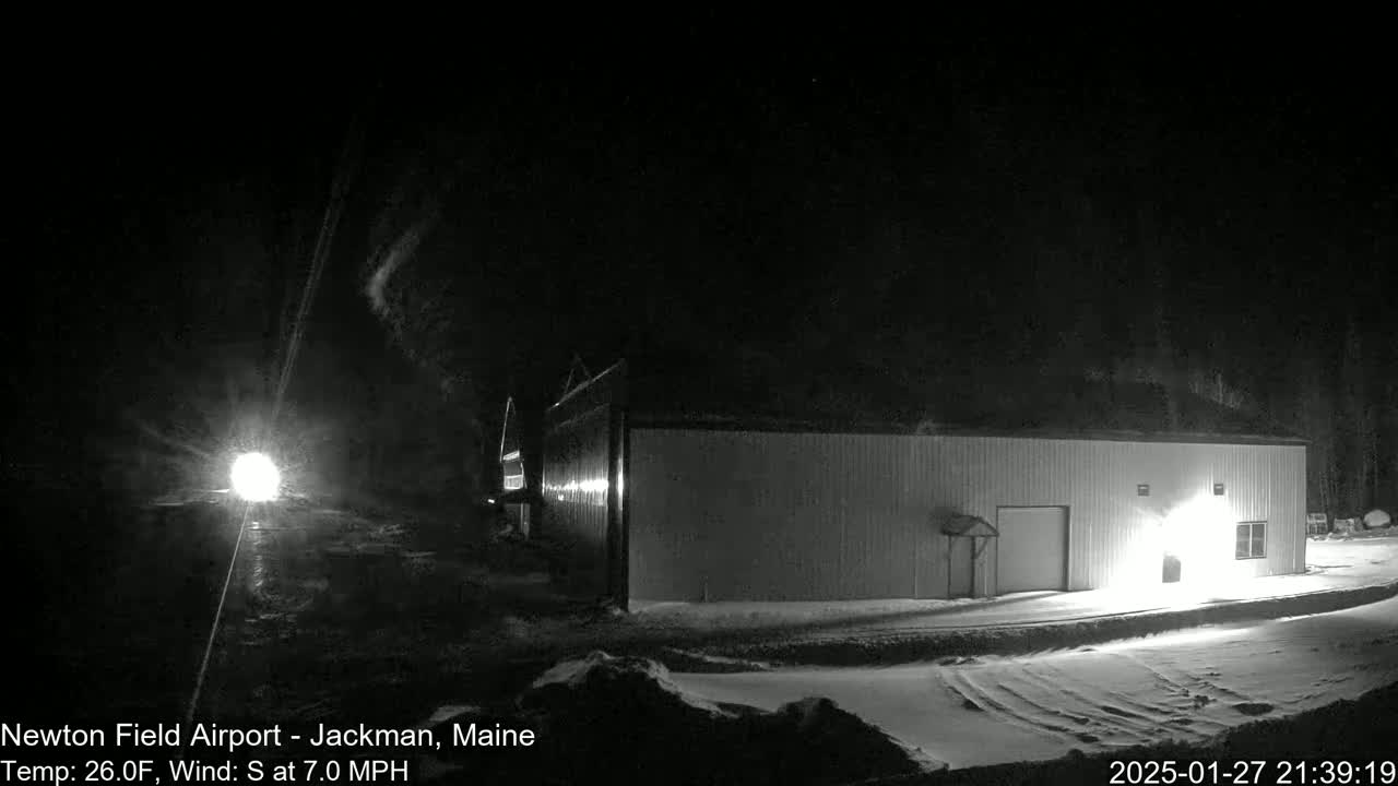 A long, low building is illuminated at night in snowy conditions with a 7 mph south wind and a temperature of 26°F.
