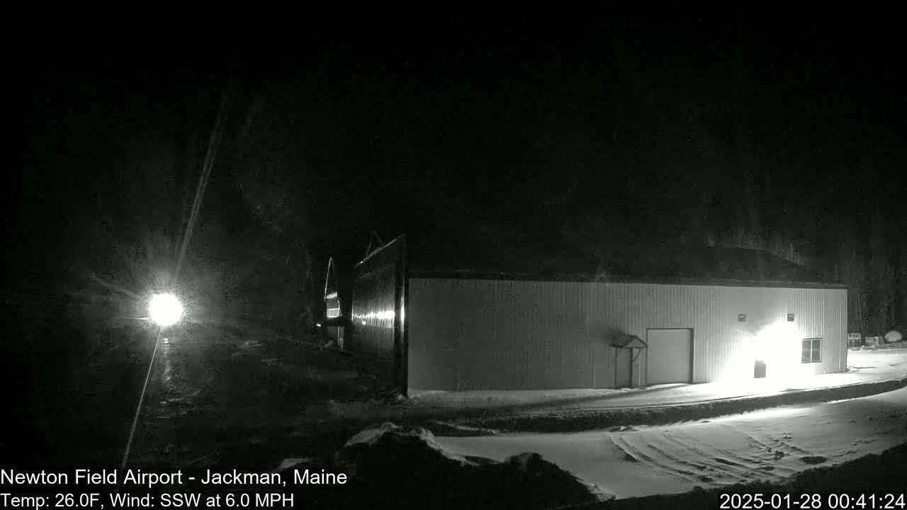 A large, light-colored building stands in a snow-covered area at night under a dark sky, illuminated by a bright light source in the distance.