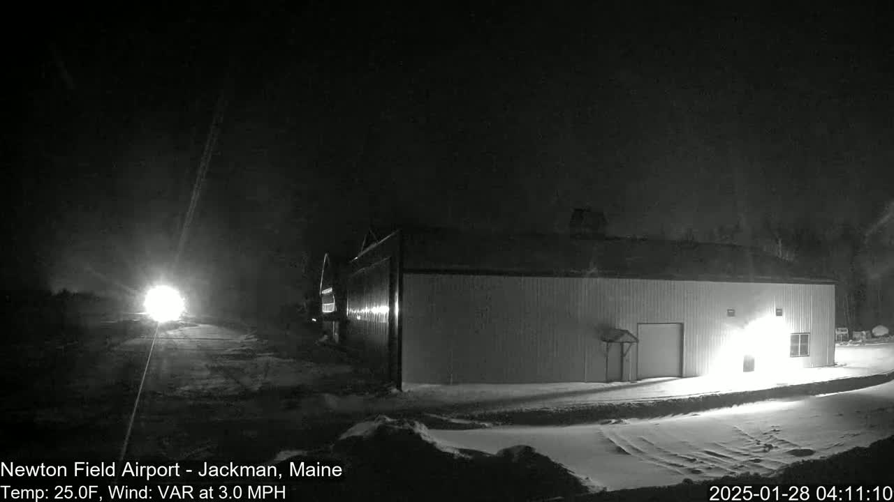 A long, low building is illuminated at night in snowy conditions, with headlights from a vehicle visible in the distance on a snow-covered road.