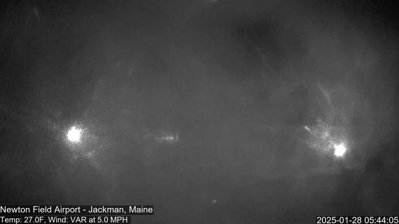 A nighttime, low-light image shows a hazy or foggy scene with two bright lights visible in the distance under a dark sky, with a 27°F temperature and 5 mph variable winds.