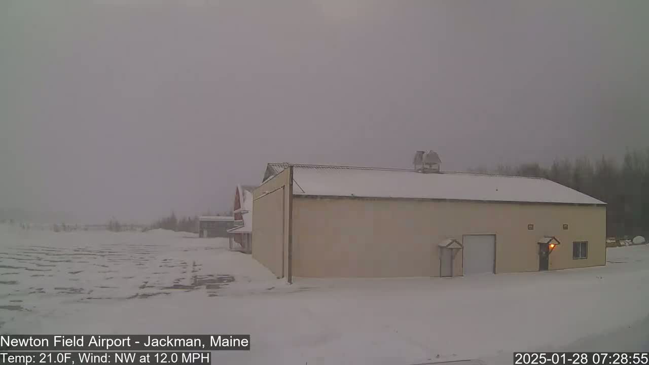 A snow-covered hangar and surrounding area are visible under an overcast sky, with a northwest wind blowing at 12 mph and a temperature of 21 degrees Fahrenheit.