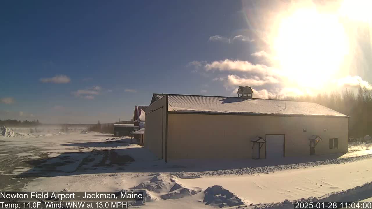 A large tan building with a snow-covered roof sits in a snowy, windy landscape under a bright, partly cloudy sun.