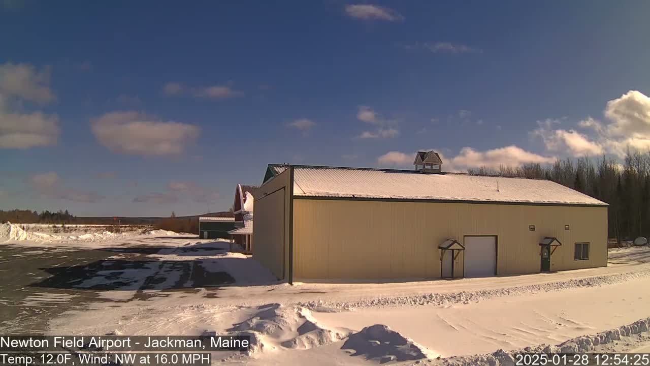 A large tan building with a snow-covered roof sits in a snowy airfield under a mostly sunny sky with some scattered clouds.