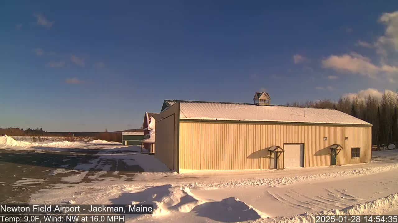 A large, tan building with a snow-covered roof sits in a snowy airfield under a mostly clear, sunny sky.