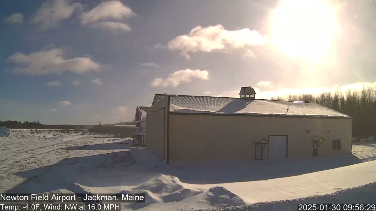 A large, light-beige building with a snow-covered roof sits in a snowy landscape under a partly sunny sky with a northwest wind.