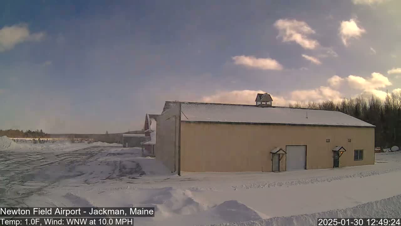 A large, tan building with a snow-covered roof stands in a snowy landscape under a partly cloudy sky.