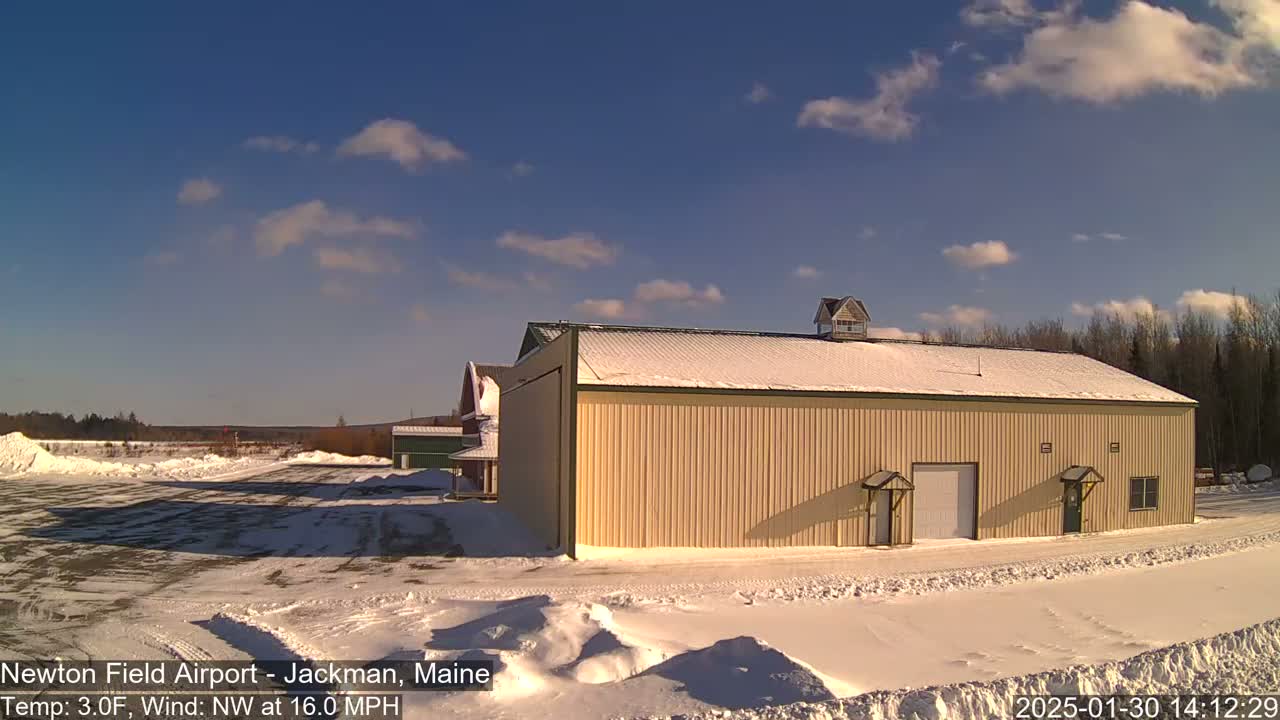A tan hangar sits on a snowy airfield under a mostly sunny sky with a few clouds.