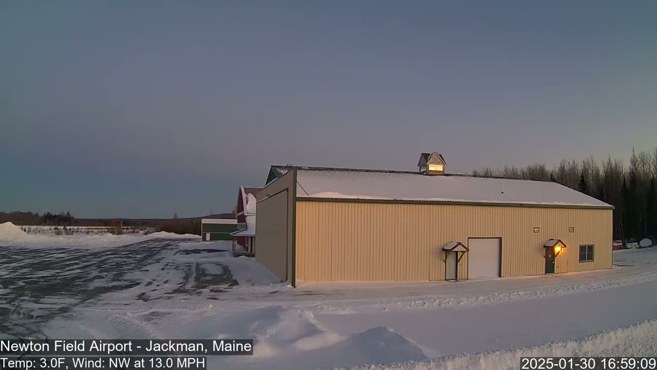 A tan hangar sits in a snow-covered airfield at dusk, with a northwest wind at 13 mph and a temperature of 3°F.