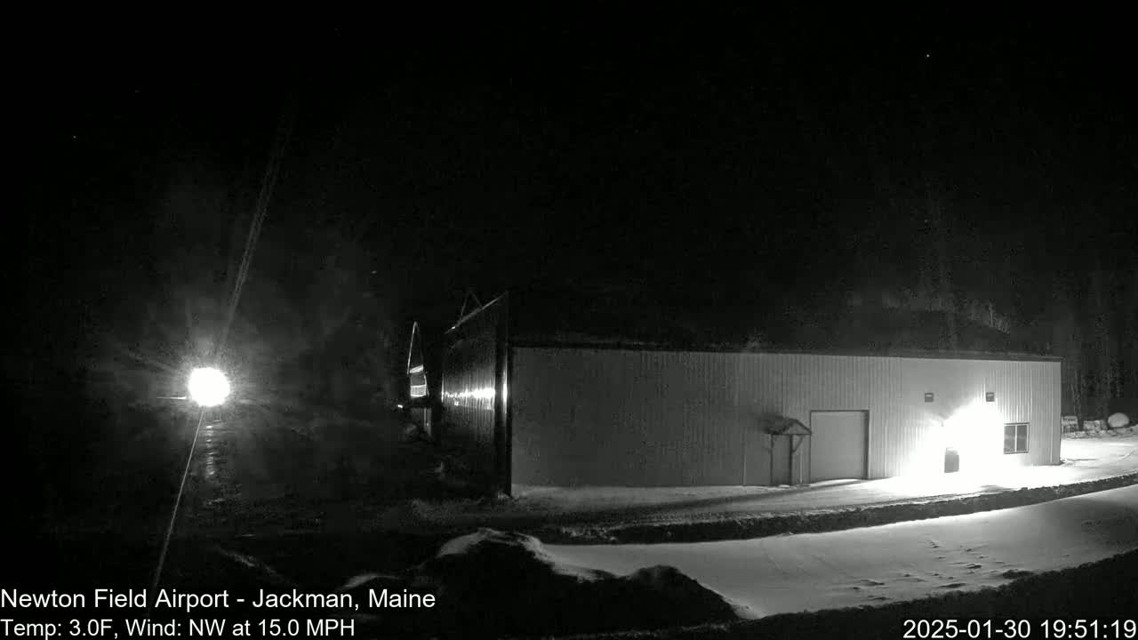 A brightly lit hangar sits in a snowy, nighttime landscape under a dark sky with a northwest wind.